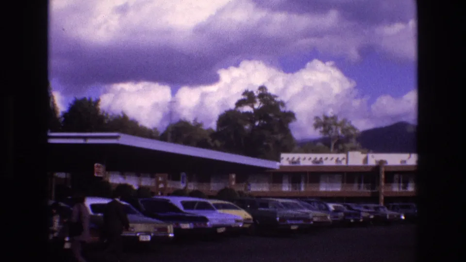 Wide 1973 New Mexico 8mm view of a low motel, a line of parked 1970s cars, and two pedestrians crossing the left foreground.