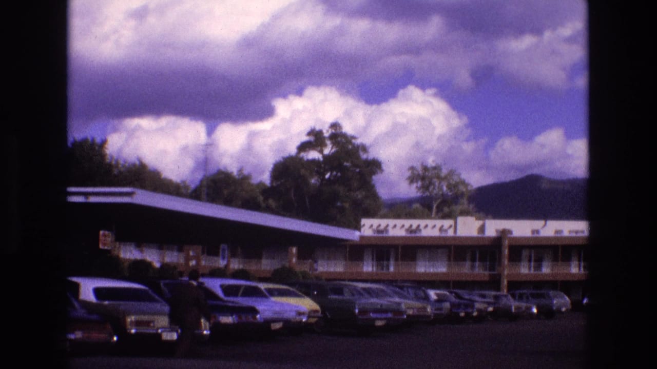 1973 New Mexico 8mm frame showing a low motel building and parking lot filled with vintage cars under dramatic clouds.