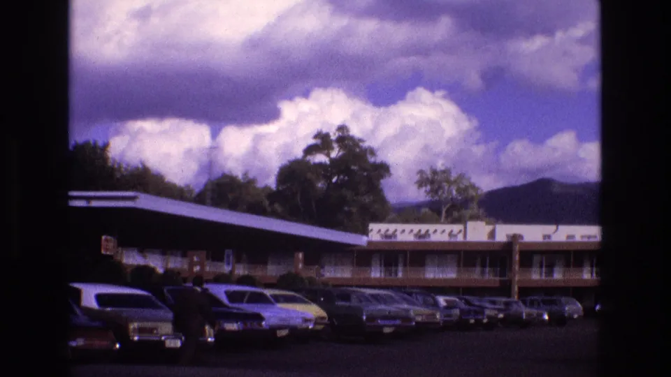 1973 New Mexico 8mm frame showing a low motel building and parking lot filled with vintage cars under dramatic clouds.