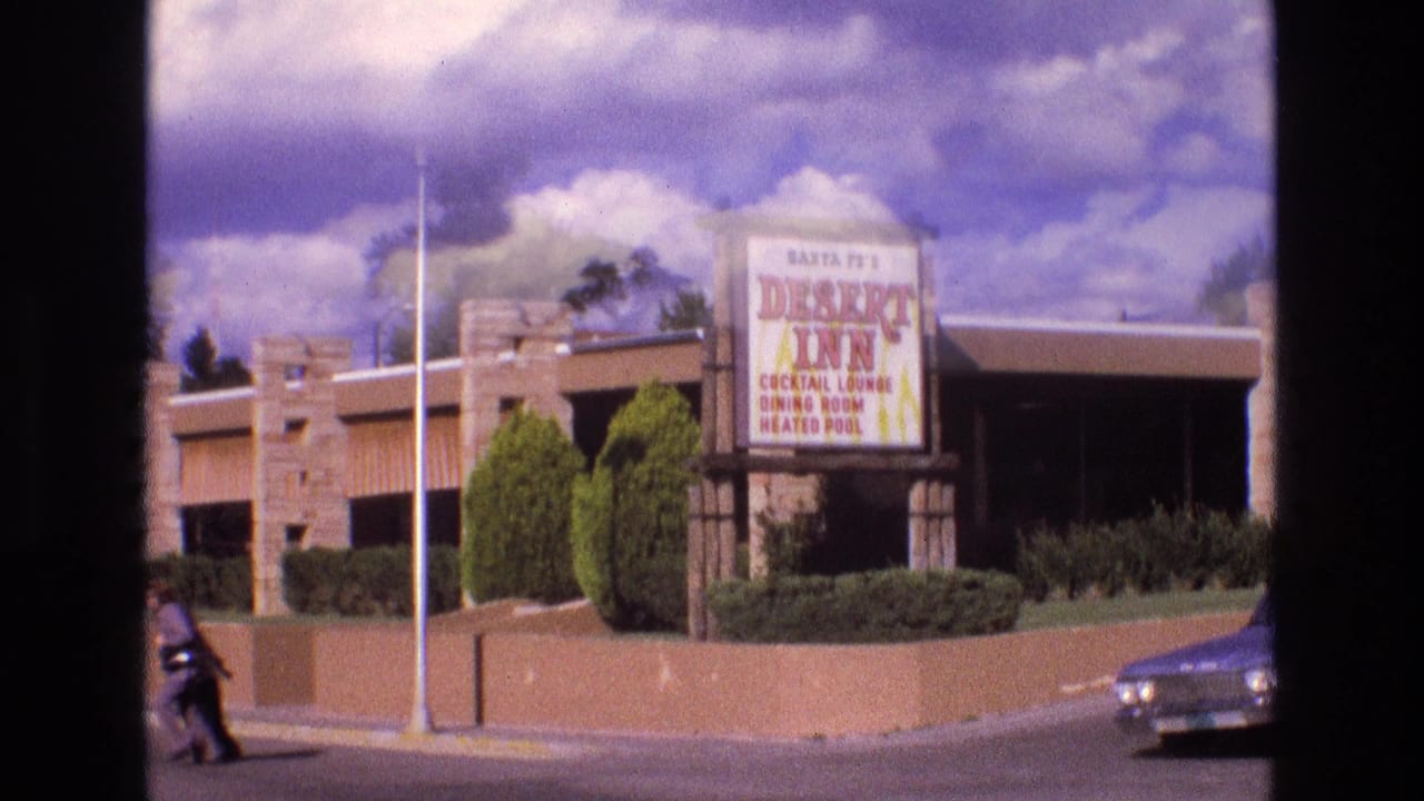 1973 New Mexico frame with the Santa Fe Desert Inn roadside sign, landscaped shrubs, a streetlight, and a car at the curb.
