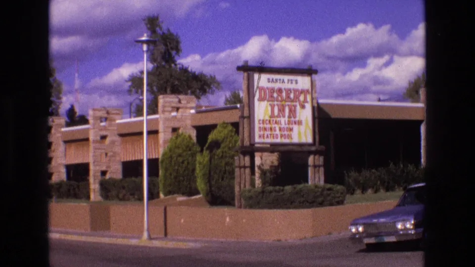 1973 New Mexico 8mm frame showing the Santa Fe Desert Inn sign, low motel building, streetlight, landscaped greenery, and a passing vintage car.
