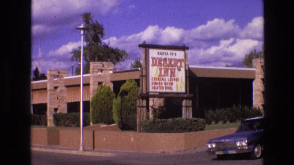 1973 New Mexico frame centered on the Santa Fe Desert Inn sign with stone-and-stucco lodging frontage, shrubs, streetlight, and a vintage car at right.