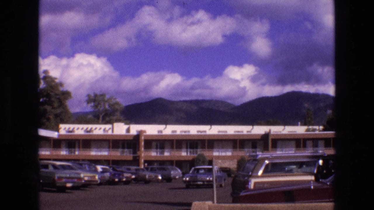 1973 New Mexico 8mm frame showing a low-rise motel or apartment-style building, a parking lot with classic cars, and distant mountains beneath a clouded sky.