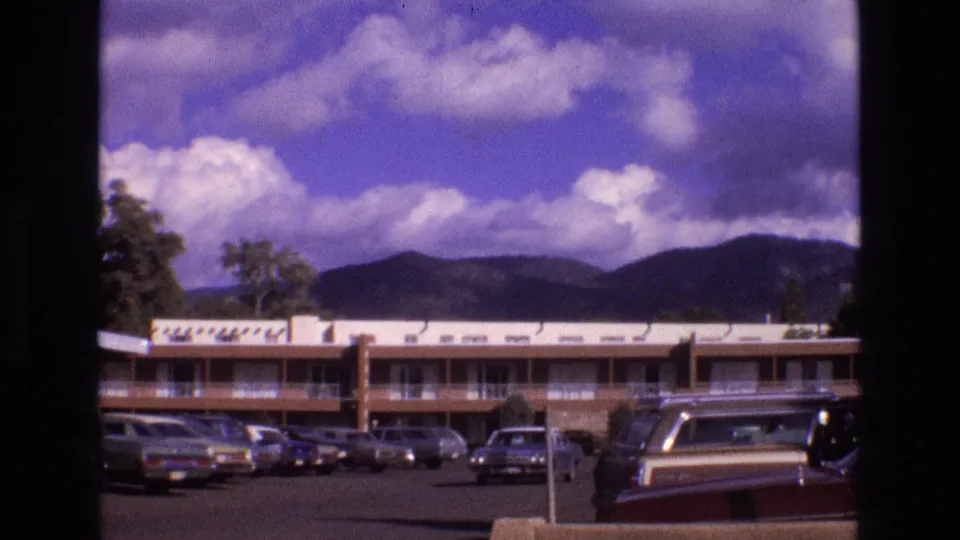 1973 New Mexico 8mm frame showing a low-rise motel or apartment-style building, a parking lot with classic cars, and distant mountains beneath a clouded sky.