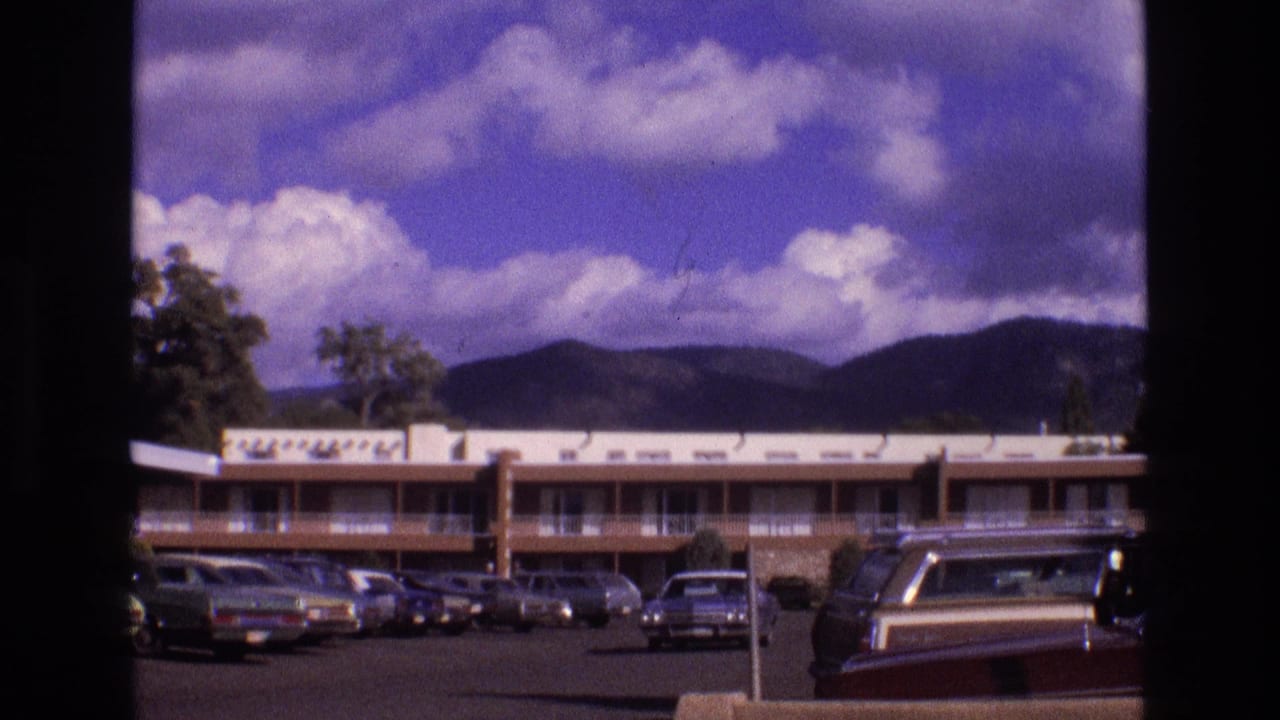1973 New Mexico home-movie frame looking through dark window or doorway framing toward a low-rise lodging building, parked cars, and a mountain skyline.