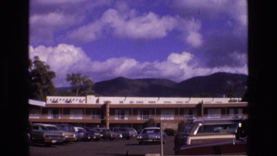 1973 New Mexico 8mm frame with a motel or apartment-style exterior, parked 1970s cars, a foreground station wagon, and distant mountains under large clouds.