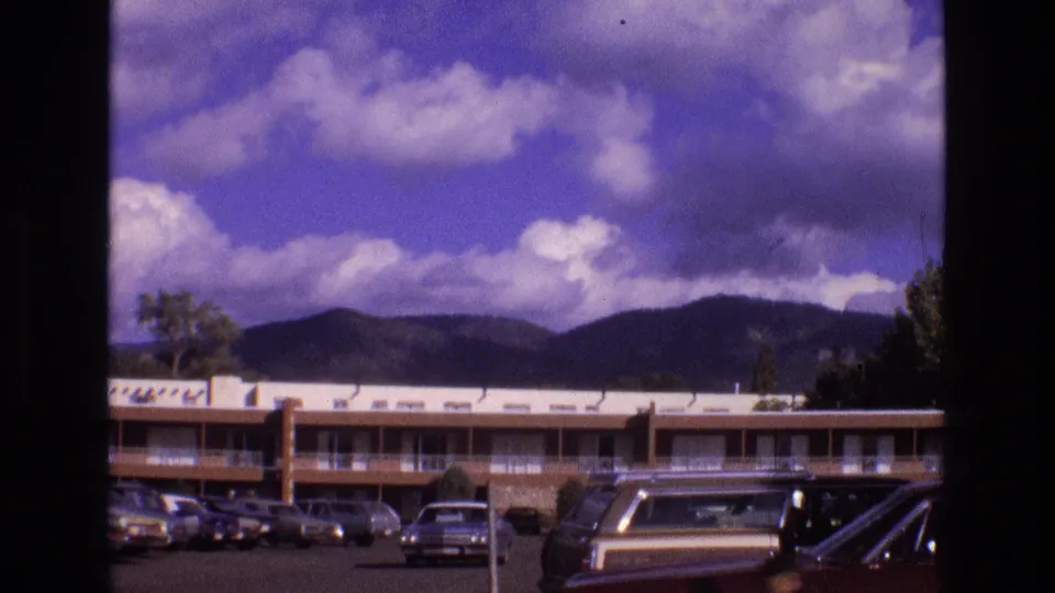 1973 New Mexico frame showing a roadside lodging exterior with parked classic cars, balcony corridors, distant mountains, and strong purple-blue 8mm color shift.