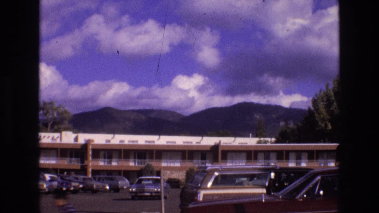 Two-story motel parking lot with parked cars, mountains, trees, and cloudy blue sky.