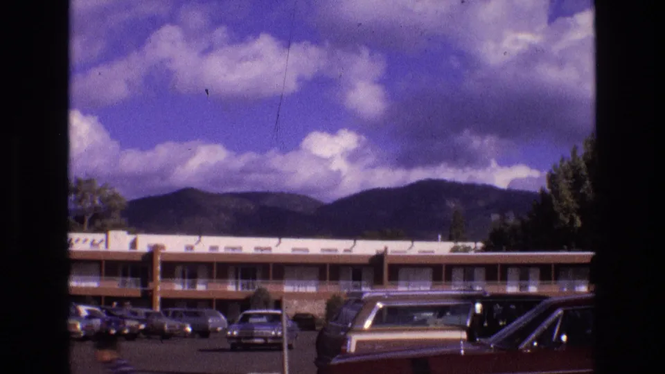 1973 New Mexico home-movie frame showing a two-story motel parking lot with older cars in front of mountains under a blue sky with scattered clouds.