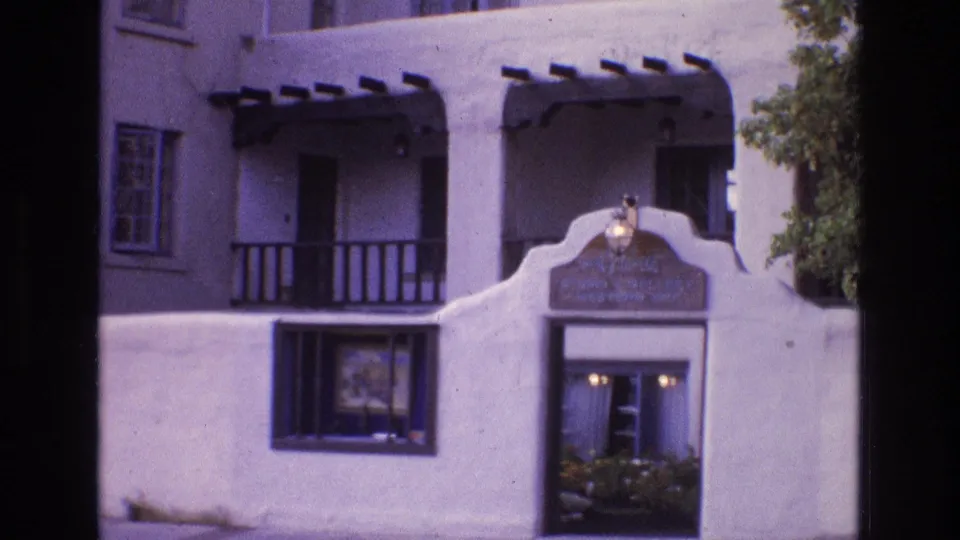 1973 New Mexico home-movie frame showing a white stucco facade with Southwestern-style wood beams, balcony rails, windows, and a decorative arched sign above an entrance.