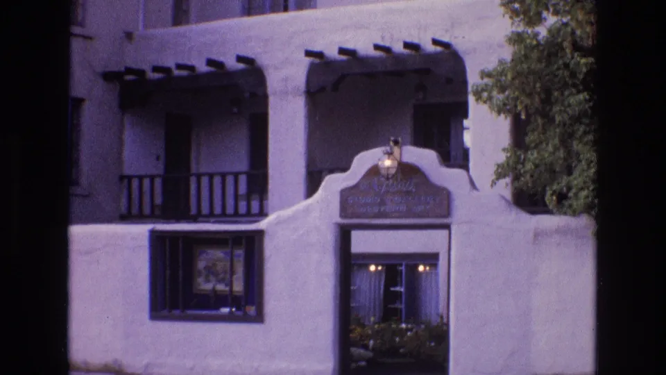 1973 New Mexico frame of a white stucco building facade with dark wood trim, balcony details, an arched sign area, windows, doorway, and a leafy tree at the side.