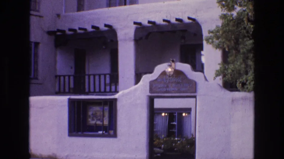 1973 New Mexico 8mm frame showing a white stucco adobe-style facade with dark wooden balcony rails, a lit doorway, windows, and a Western art gallery sign.