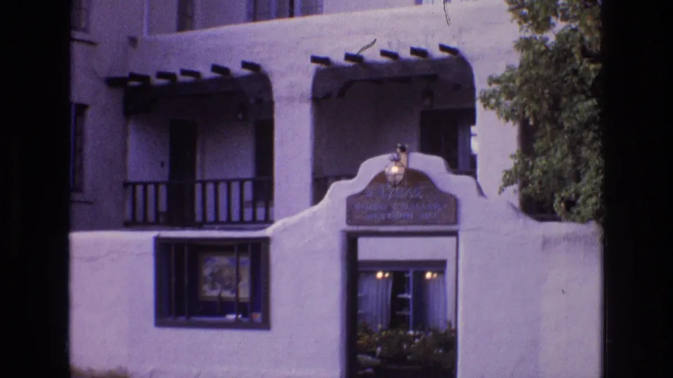 1973 New Mexico 8mm frame of a Southwestern stucco building entrance with curved entry wall, dark balcony rails, doorway lights, and tree foliage at the right edge.