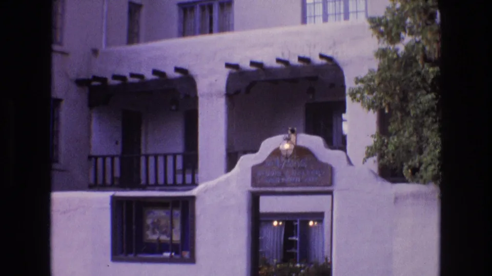 1973 New Mexico 8mm frame showing the gallery entrance more tightly, with white stucco walls, wooden balcony rails, a barred window, and warm lights inside the doorway.
