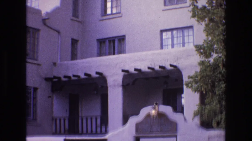 1973 New Mexico 8mm frame with the camera shifted right, showing upper windows, dark wooden balcony rails, a curved stucco entry wall, the gallery sign, and tree foliage.