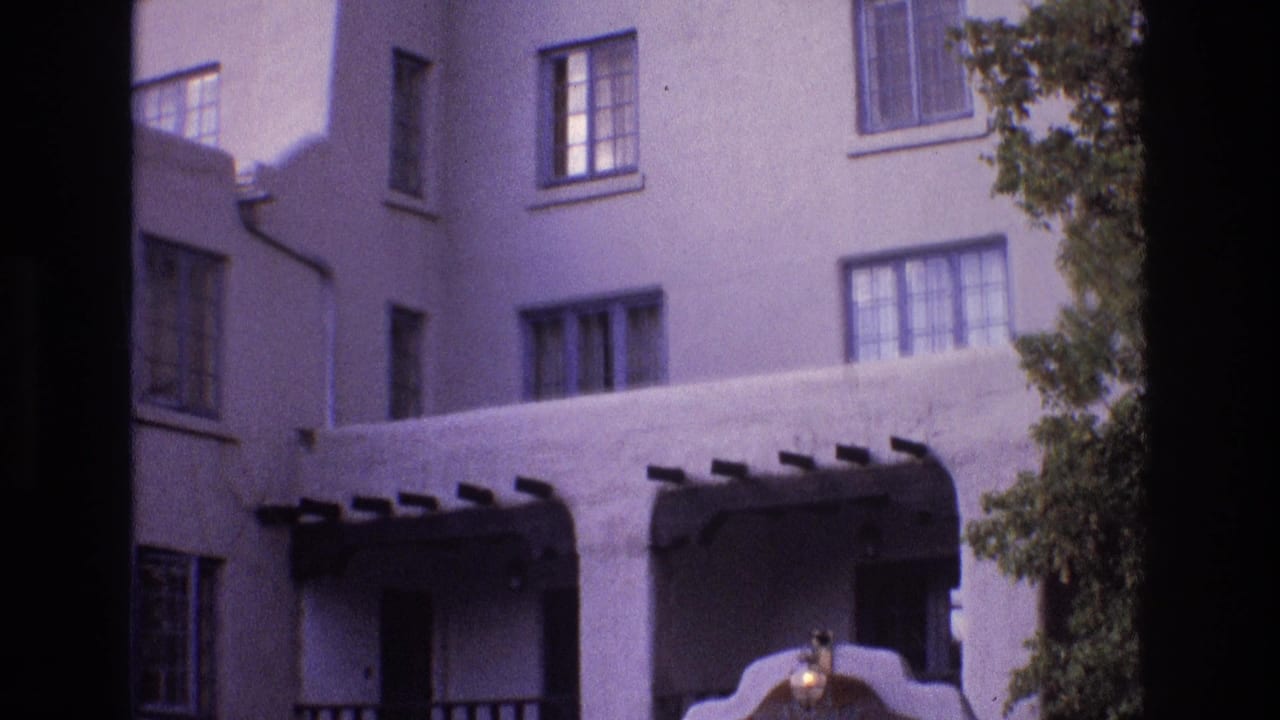 1973 New Mexico 8mm frame showing the upper portion of a white stucco building with rectangular windows, balcony openings, dark projecting beams, and foliage along the side.