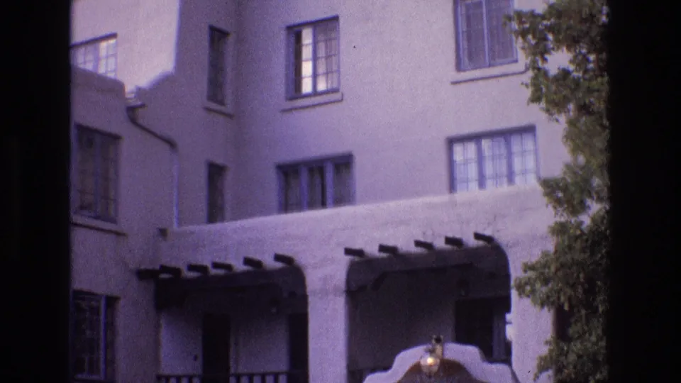 1973 New Mexico 8mm frame showing the upper portion of a white stucco building with rectangular windows, balcony openings, dark projecting beams, and foliage along the side.