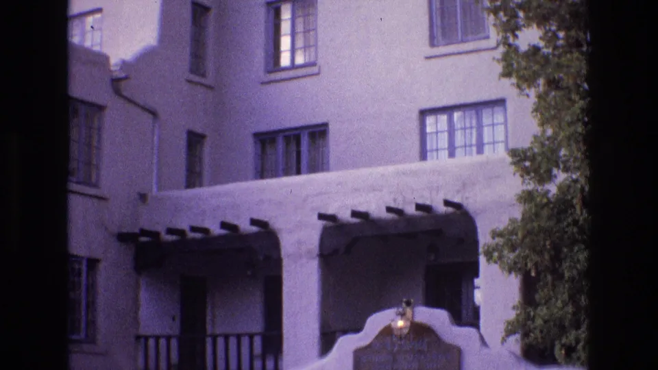 1973 New Mexico 8mm frame of a stucco building exterior with upper windows, shaded balcony recesses, dark beams, a partial curved entrance feature, and tree foliage.