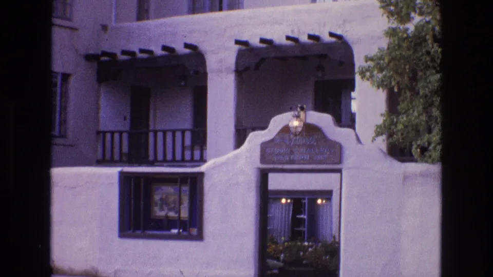 1973 New Mexico 8mm frame showing the lower building facade with a distinctive curved stucco entrance, illuminated sign or plaque area, balcony railings, window display, and dark beam accents.