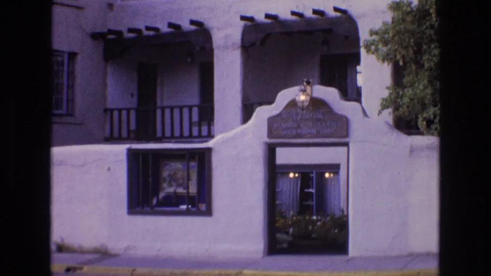 1973 New Mexico 8mm frame showing a sunlit white stucco adobe-style facade with dark wooden balconies, a curved entrance wall, and a window facing the street.