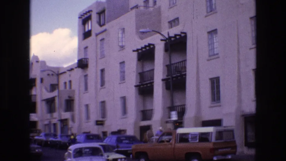 1973 New Mexico 8mm frame looking down a street lined with white stucco buildings, parked mid-century cars, a brown pickup truck, balconies, and sidewalk activity.