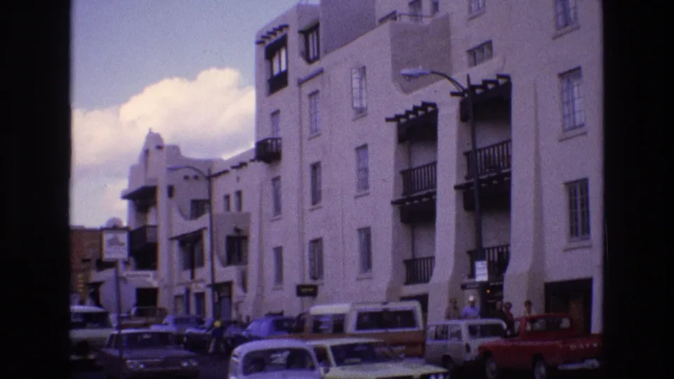 1973 New Mexico street frame with adobe-style stucco facades, balcony shadows, parked cars, a pickup truck, street signs, and pedestrians near the curb.