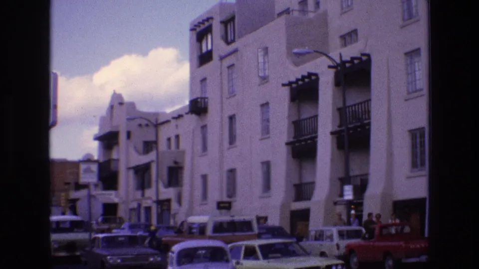 1973 New Mexico home-movie frame showing a white stucco multi-story building with dark balconies, parked cars, pedestrians, and street traffic in front.