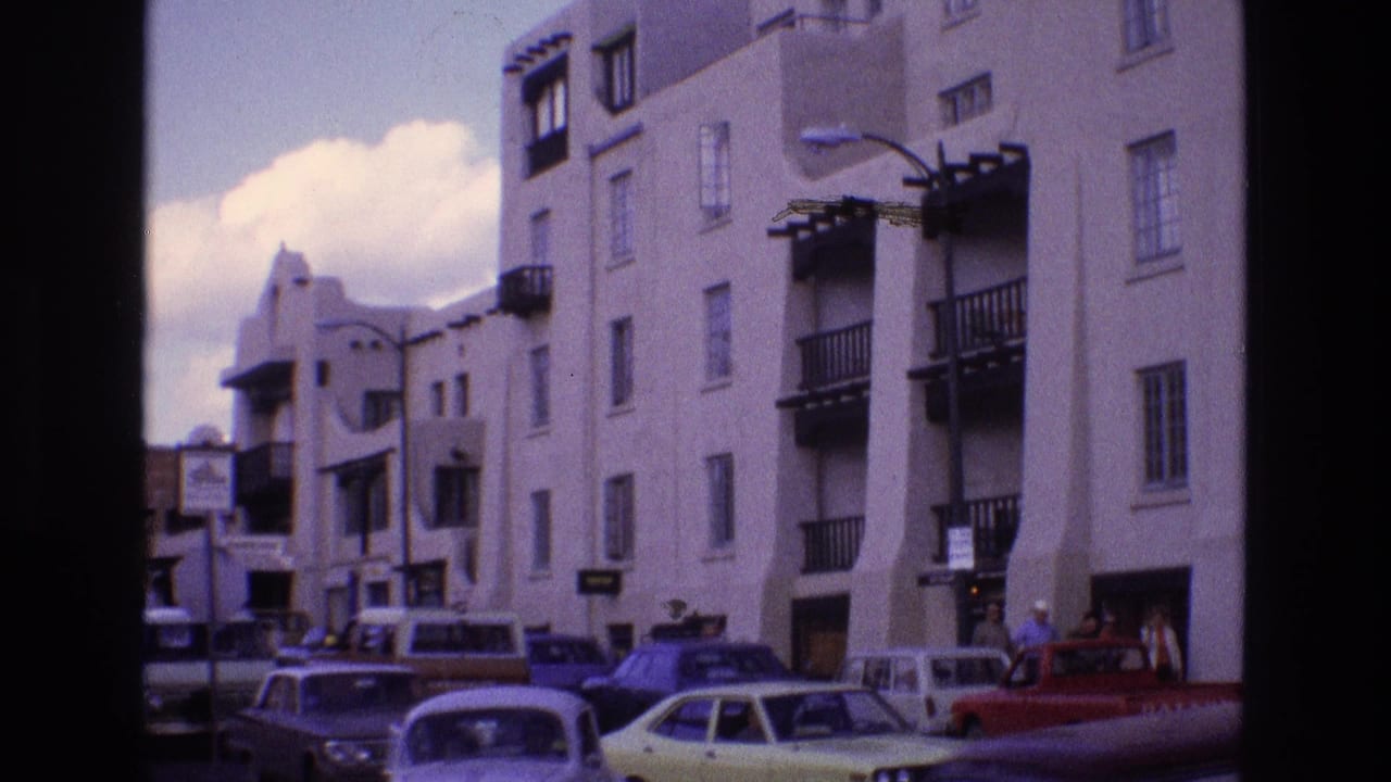 1973 New Mexico street view with older American cars, a red pickup, pedestrians, street signage, and a white stucco building with balconies.