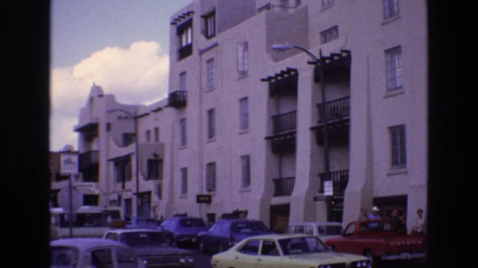 1973 New Mexico home-movie frame showing crowded curbside traffic, a pale car, a red pickup, pedestrians, and the white stucco building front.