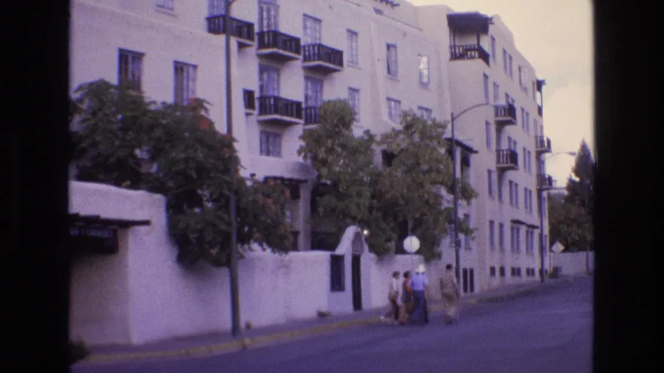 1973 New Mexico frame showing the side of a white stucco multi-story building, balconies, trees, a low wall, streetlamp, and pedestrians on a quieter street.