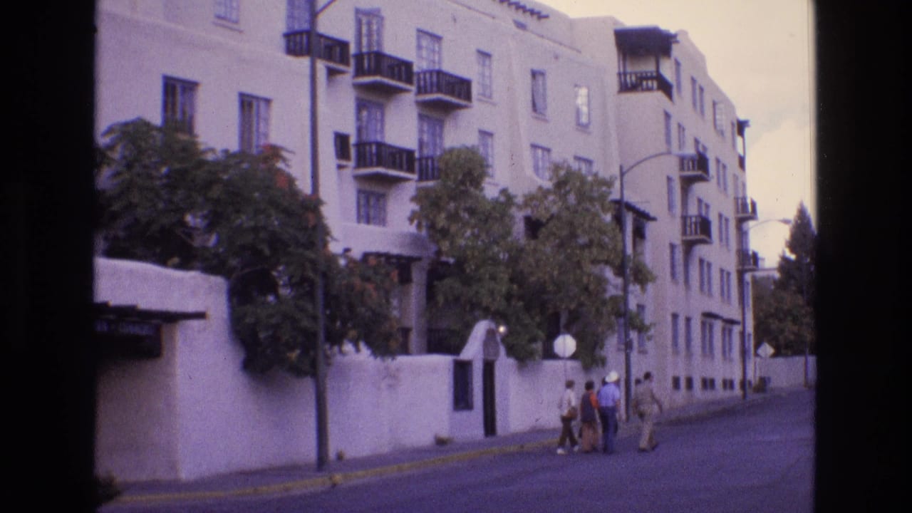 1973 New Mexico home-movie frame showing a white stucco apartment building with black balconies, trees, a low wall, and several people walking along the sidewalk.