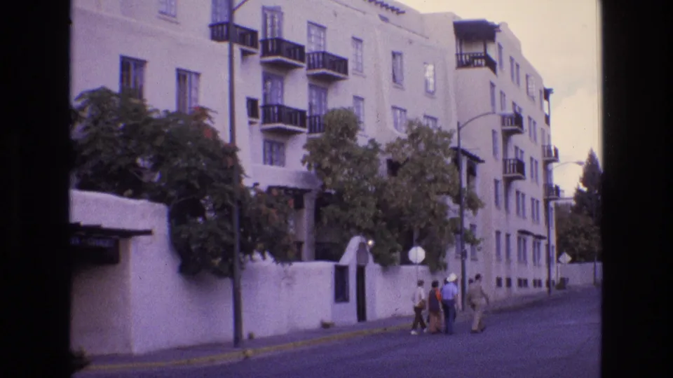 1973 New Mexico home-movie frame showing a white stucco apartment building with black balconies, trees, a low wall, and several people walking along the sidewalk.
