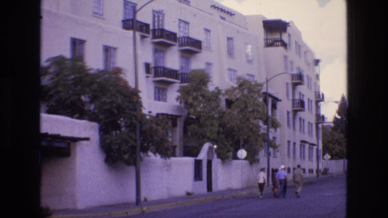 1973 New Mexico frame of a residential apartment exterior with black balconies, a white perimeter wall, trees, and a small group walking down the sidewalk.