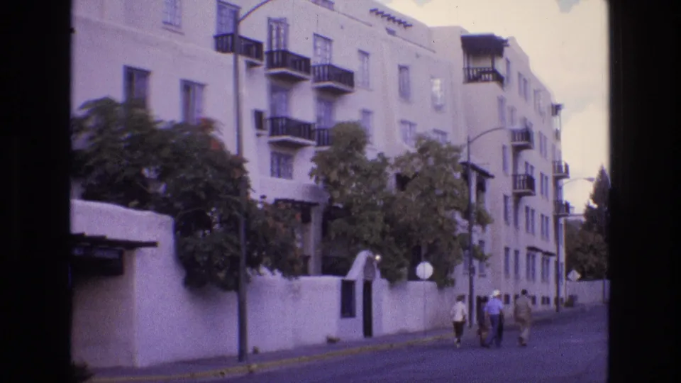 1973 New Mexico frame of a residential apartment exterior with black balconies, a white perimeter wall, trees, and a small group walking down the sidewalk.