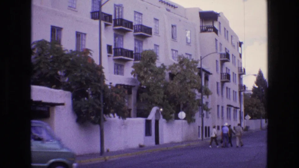 1973 New Mexico home-movie frame with pedestrians walking past a white stucco apartment building, black balconies, trees, and a low wall along the street.