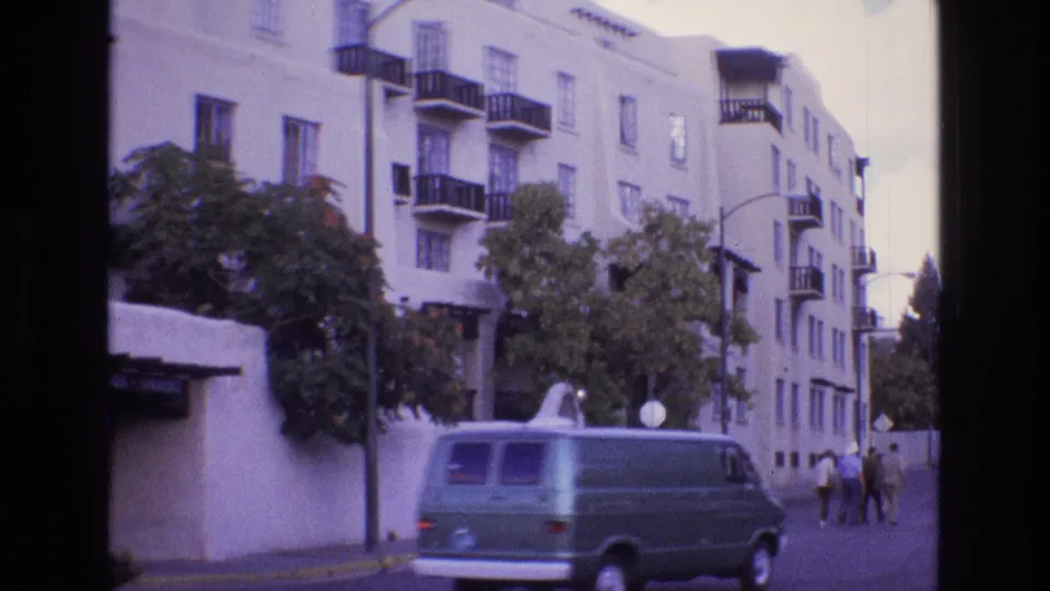1973 New Mexico frame showing a green van driving past a white stucco apartment building with balconies as pedestrians continue along the sidewalk.