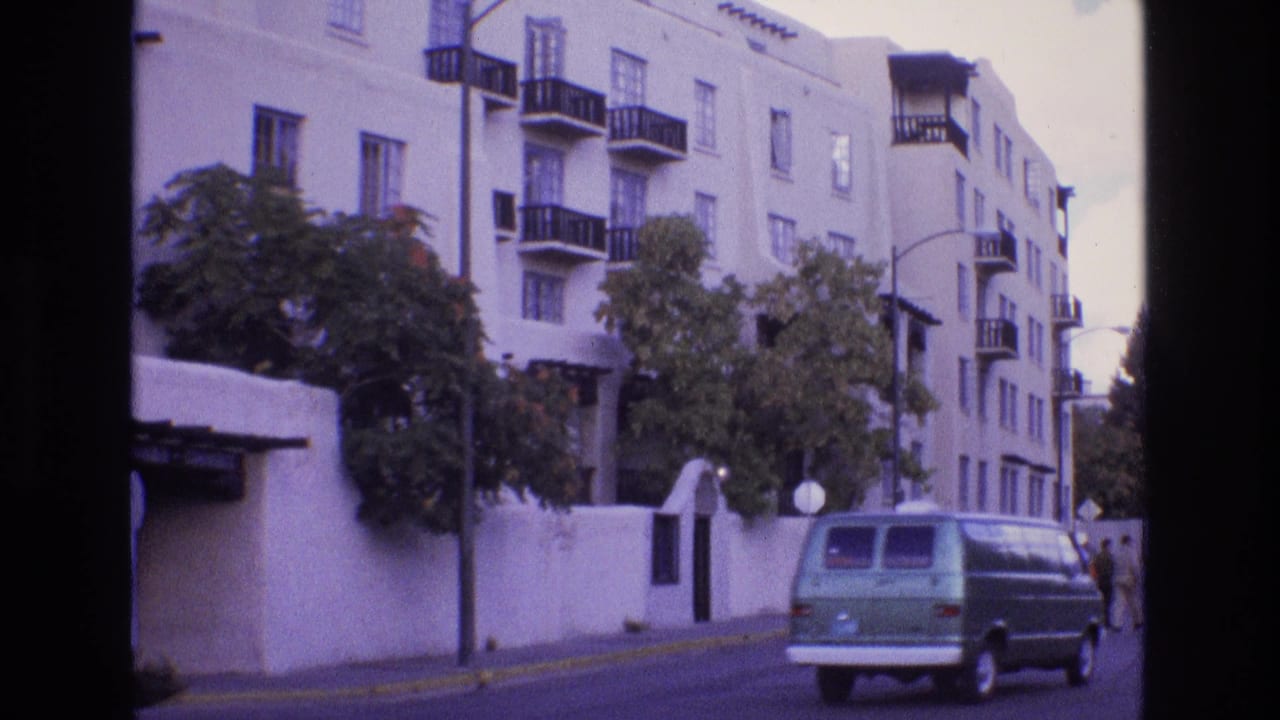 1973 New Mexico home-movie frame showing a green van driving past a white stucco apartment building with dark balcony railings, street trees, and distant pedestrians.