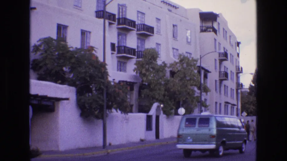 1973 New Mexico home-movie frame showing a green van driving past a white stucco apartment building with dark balcony railings, street trees, and distant pedestrians.