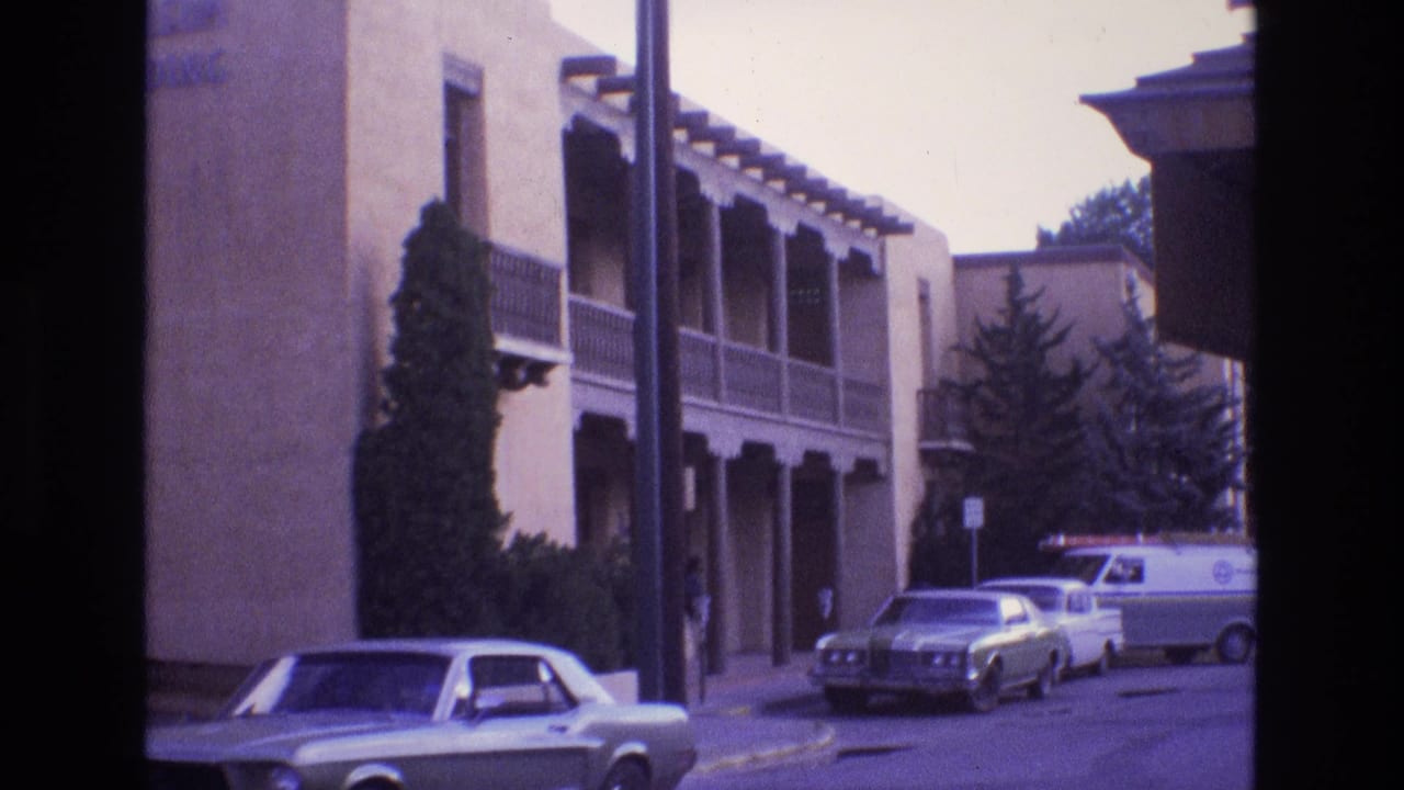 Residential street with adobe-style building, parked cars, and a white van near the curb.