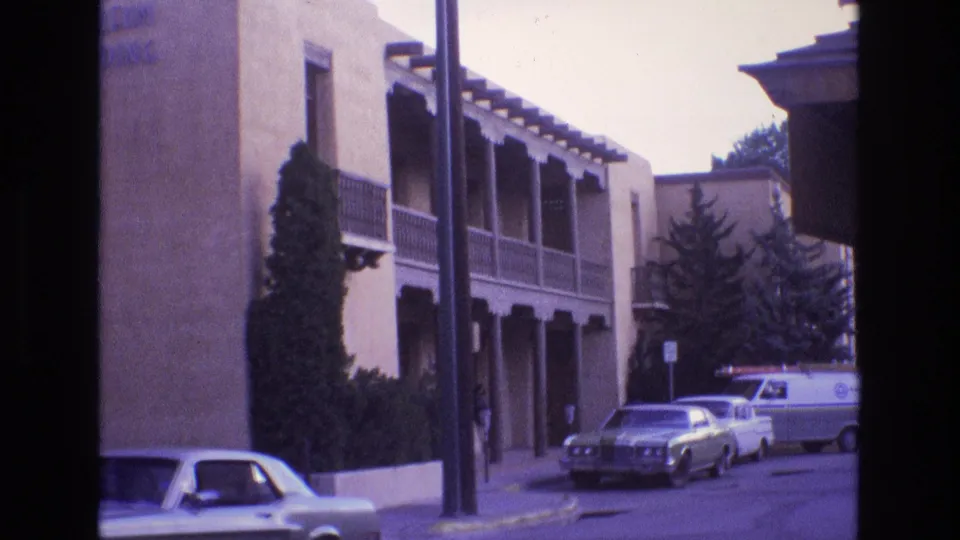 1973 New Mexico home-movie frame looking along a quiet residential street with an adobe-style building, wooden balcony supports, parked cars, and a white van.