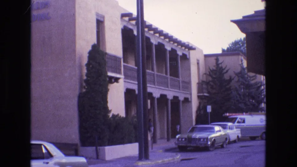 1973 New Mexico street frame showing parked vehicles outside an adobe-style apartment building with wooden balconies, a pole at the curb, and trees in the background.