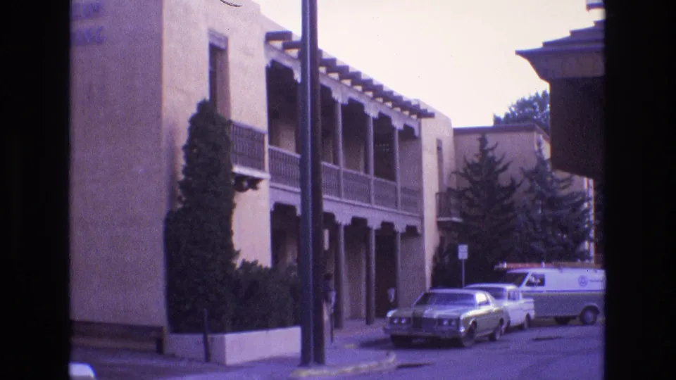 1973 New Mexico 8mm frame showing a quiet street beside a stucco building with balcony supports, landscaping, parked cars, and a white van.