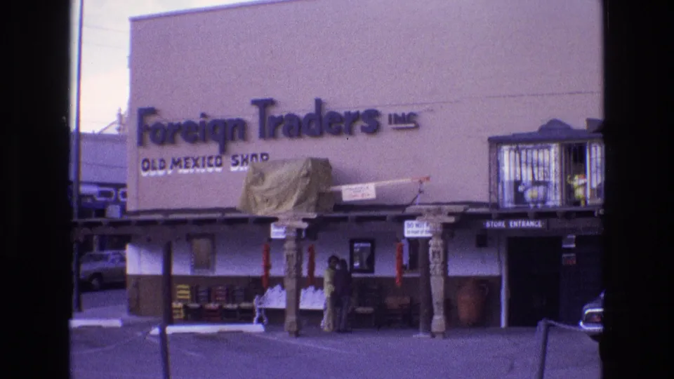 1973 New Mexico storefront frame showing the Foreign Traders Inc. Old Mexico Shop sign, entrance area, outdoor display items, parked vehicles, and people near the building.