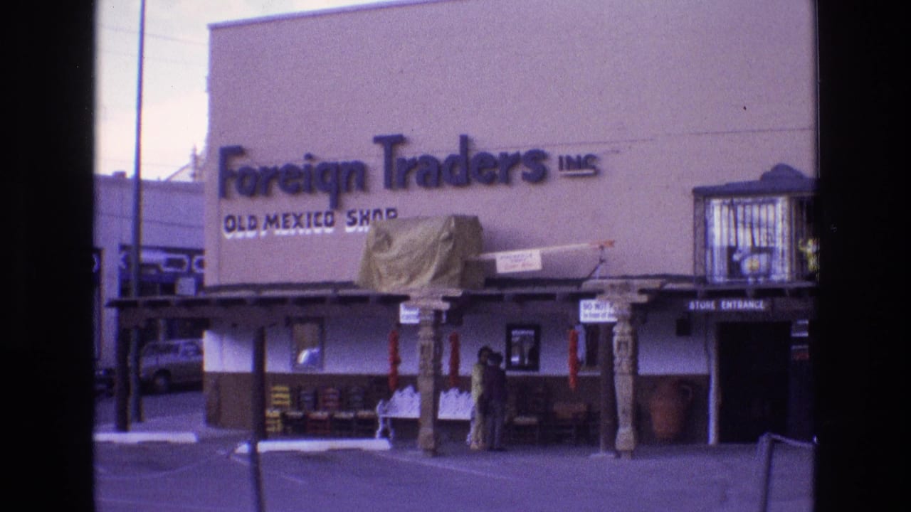 1973 New Mexico 8mm frame showing the Foreign Traders Inc. Old Mexico Shop exterior, with a covered storefront, carved posts, displayed goods, two people near the door, and a parked car at the side.