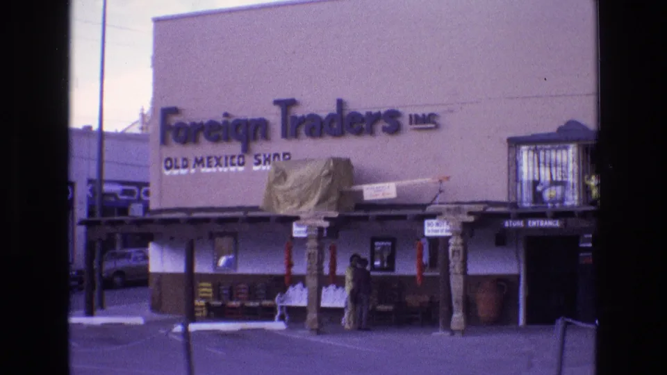 1973 New Mexico 8mm frame showing the Foreign Traders Inc. Old Mexico Shop exterior, with a covered storefront, carved posts, displayed goods, two people near the door, and a parked car at the side.