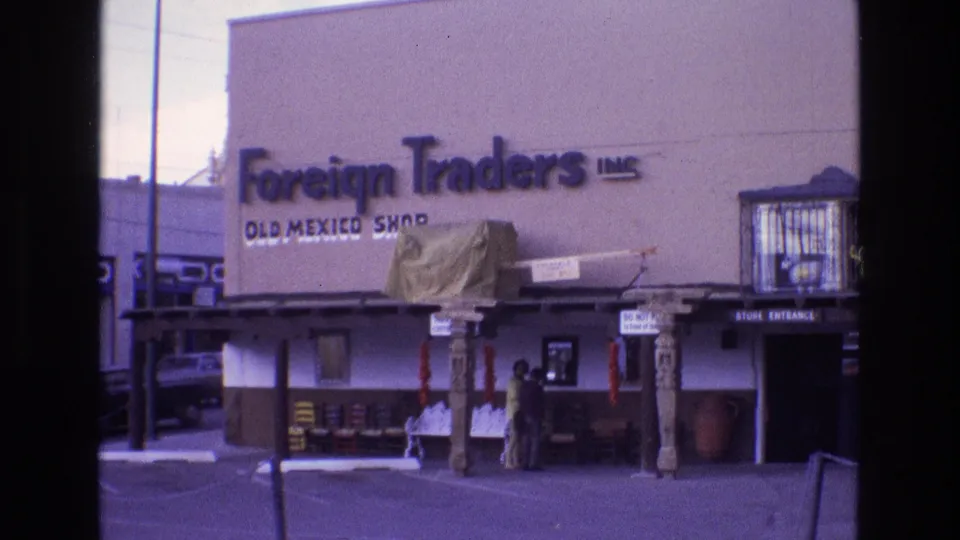 1973 New Mexico 8mm frame holding on the Foreign Traders Inc. Old Mexico Shop exterior, with a stucco facade, covered walkway, carved supports, outdoor merchandise, two people by the entrance, and a parked car partly visible to the left.
