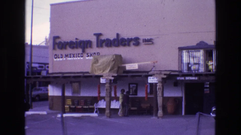1973 New Mexico frame from an 8mm home movie showing the Foreign Traders Inc. Old Mexico Shop building, including the large facade sign, dark store entrance, decorative posts, exterior displays, and two visible people near the front.
