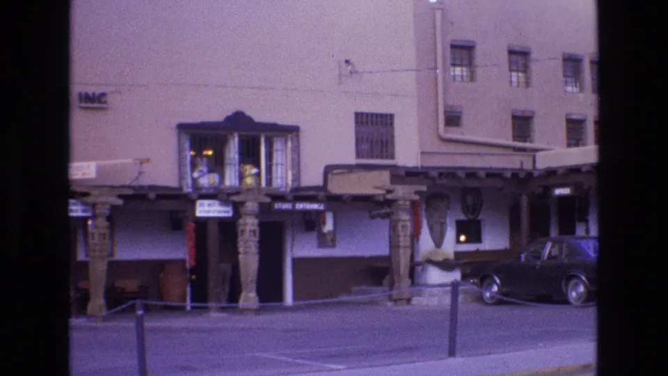 1973 New Mexico street scene showing an adobe-style business frontage with carved wooden columns, storefront signs, and a dark sedan parked in front.