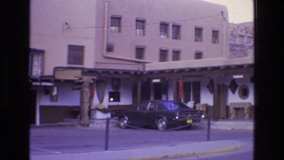 A 1973 New Mexico town business exterior with a dark sedan, yellow rear plate, carved support posts, and adobe-style upper walls visible from across the street.
