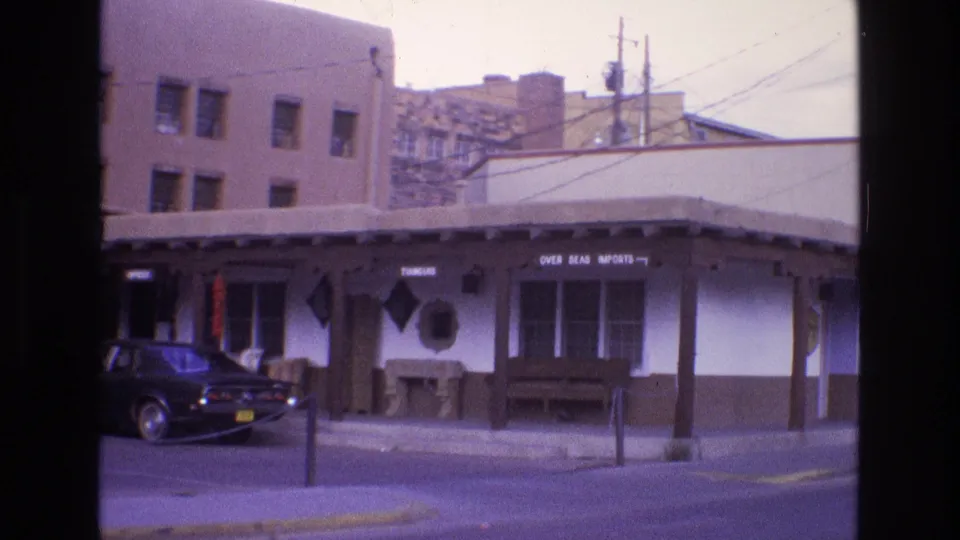1973 New Mexico home-movie frame showing the storefront entrance area, signs above the covered walkway, a stone fireplace-like feature, and the rear of a parked car at left.